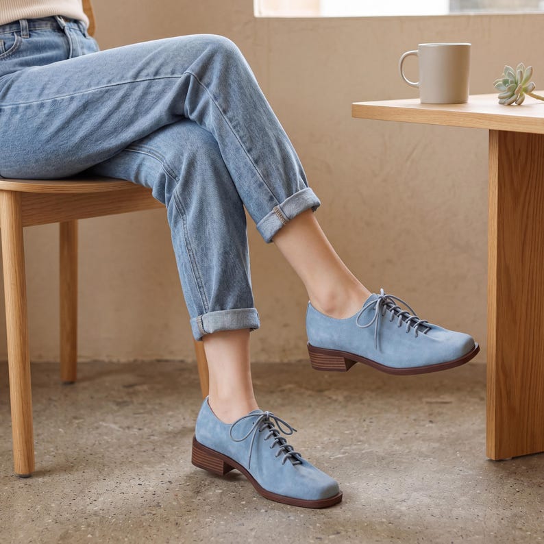 May include: Light blue suede oxford shoes with brown heels and laces. The shoes are worn by a person wearing blue jeans. The shoes have a classic design and a low heel.