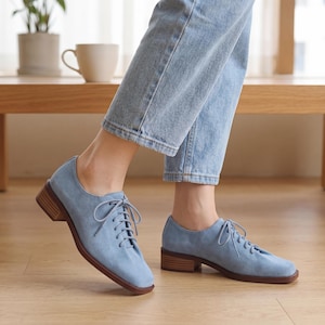 May include: Light blue suede oxford shoes with a lace-up design and a low, stacked heel. The shoes have a rounded toe and a brown sole. The shoes are worn with light blue jeans.