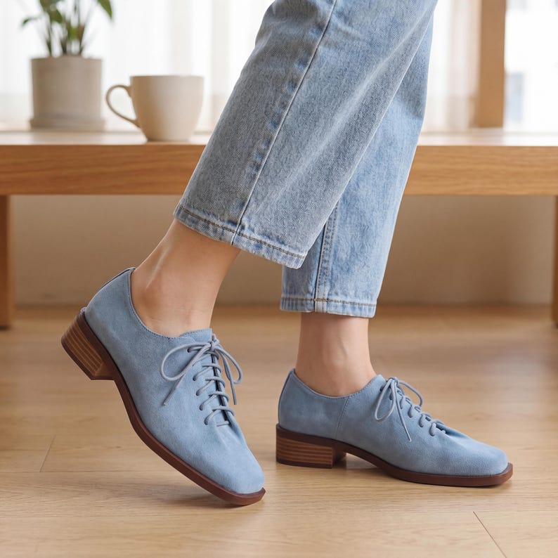 May include: Light blue suede oxford shoes with a lace-up design and a low, stacked heel. The shoes have a rounded toe and a brown sole. The shoes are worn with light blue jeans.