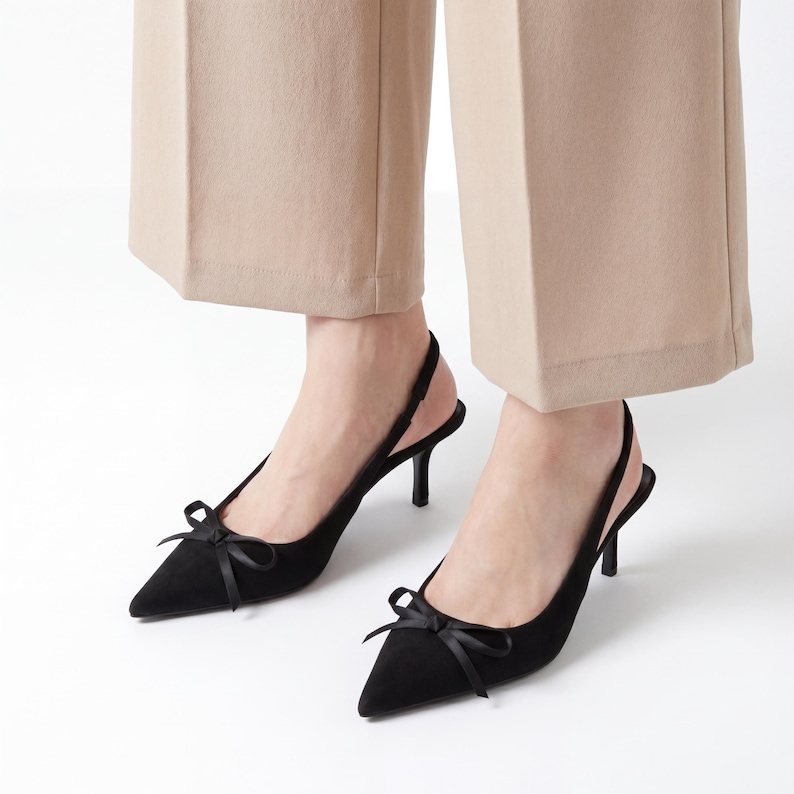 May include: Black slingback heels with pointed toes and small bows. The heels are approximately 3 inches tall. The shoes are made of a soft, suede-like material. The background is white.