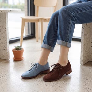 May include: A pair of suede oxford shoes, one light blue and one brown, with matching laces and low heels. The shoes are worn with blue jeans. The shoes are a stylish choice for everyday wear.