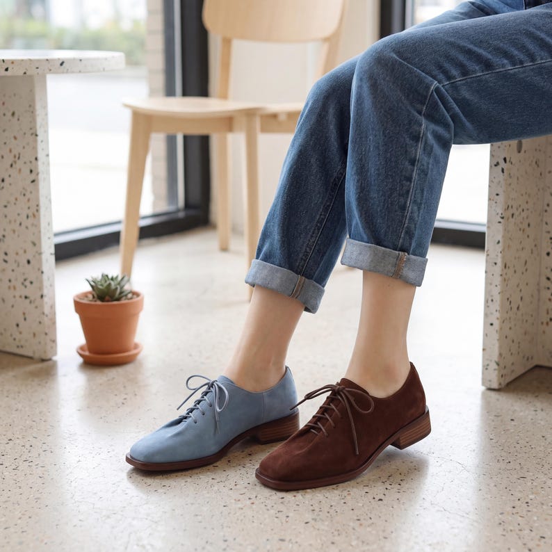May include: A pair of suede oxford shoes, one light blue and one brown, with matching laces and low heels. The shoes are worn with blue jeans. The shoes are a stylish choice for everyday wear.