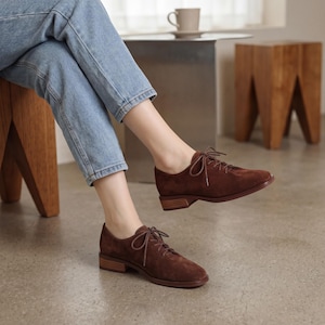 May include: Brown suede lace-up shoes with a low heel. The shoes have a classic Oxford style and are worn with blue jeans. The shoes are a warm brown color and have a simple, elegant design. The heels are approximately 1 inch high.