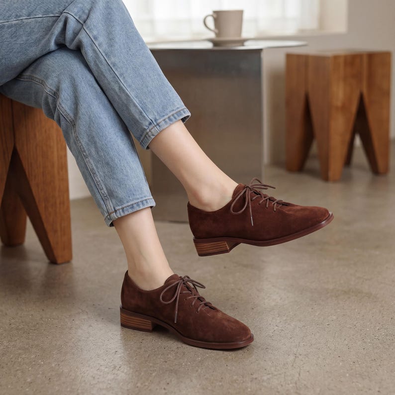 May include: Brown suede lace-up shoes with a low heel. The shoes have a classic Oxford style and are worn with blue jeans. The shoes are a warm brown color and have a simple, elegant design. The heels are approximately 1 inch high.