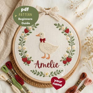 May include: An embroidery hoop containing a cream fabric with a stitched white duck wearing a red bow, surrounded by a wreath of green leaves, white flowers, and red strawberries. The name Amelie is embroidered in red script below the duck.