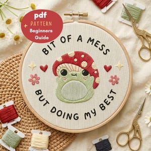 May include: An embroidery hoop containing a fabric piece with a stitched design of a green frog wearing a red mushroom cap. The text reads BIT OF A MESS BUT DOING MY BEST. The design includes small pink flowers, red hearts, and yellow stars.