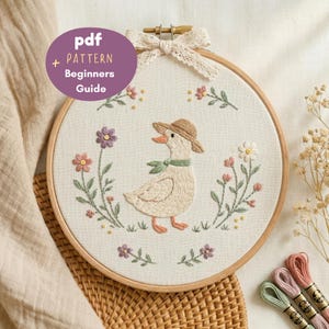 May include: A round wooden embroidery hoop containing white fabric with a hand-stitched design of a duck wearing a straw hat and a green scarf, surrounded by floral stems. A purple circular overlay displays the text pdf + PATTERN Beginners Guide.