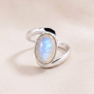 May include: A silver ring featuring a smooth, oval-shaped moonstone cabochon set in a polished metal band. The stone displays a soft, iridescent blue glow against the metallic surface, with the band curving around the central gemstone setting.