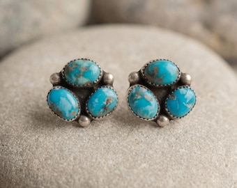 May include: A pair of stud earrings featuring three oval-shaped turquoise stones set in silver. Each earring is arranged in a cluster formation with small silver beads placed between the stones, showcasing natural blue and brown veining patterns.