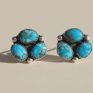 May include: A pair of silver-toned stud earrings, each featuring three oval turquoise stones. The earrings have a cluster design with a small silver ball between each turquoise stone. The turquoise stones have a mottled appearance.