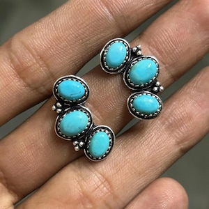 May include: Two pairs of turquoise stud earrings. Each earring features three oval turquoise stones set in a silver-colored metal. The earrings are displayed on a hand, showcasing the bright blue color of the stones.