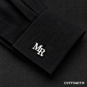 May include: A silver-toned metal cufflink featuring the letters MR in a serif font, attached to a black fabric shirt cuff with visible stitching. The cufflink is positioned on the corner of the folded cuff, which has a fine, textured surface.