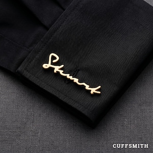 May include: Close-up of a black dress shirt cuff with a gold-colored cursive "Stuart" cufflink. The cufflink is a stylish accessory. The word "CUFFSMITH" is printed in white at the bottom right.