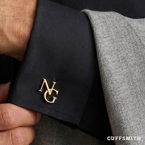 May include: A gold-toned metal cufflink featuring the letters NG in a serif font, attached to the black fabric cuff of a long-sleeved shirt. The cuff is positioned next to a textured grey fabric sleeve, with the text CUFFSMITH visible at the bottom.