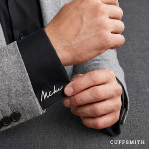 May include: Silver cufflinks with the handwritten word "Mcke" on a black shirt cuff. The cuff is part of a gray suit jacket. The word "CUFFSMITH" is printed in white at the bottom right corner.