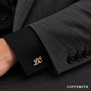 Op de afbeelding: Een goudkleurige metalen manchetknop met de cursieve letters LC, bevestigd aan de zwarte manchet van een overhemd met lange mouwen, gedragen onder een donkergrijze, gestructureerde blazer met drie zwarte knopen op de mouw en de tekst CUFFSMITH in de hoek.
