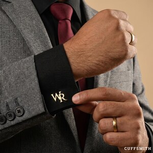Op de afbeelding: Een goudkleurige manchetknop met de letters WR in een schreeflettertype, bevestigd aan een zwarte overhemdmanchet. De manchet wordt gedragen onder een grijze, gestructureerde blazer met een donkerrode das om de pols.