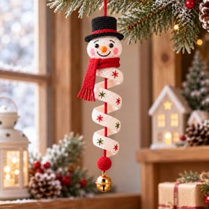 Op de afbeelding: Een gehaakte sneeuwpop-ornament met een zwarte hoge hoed, een rode sjaal en een wit spiraalvormig lichaam versierd met kleine rode en groene sterpatronen. Het ornament hangt aan een rood koord en eindigt in een kleine goudkleurige metalen bel.