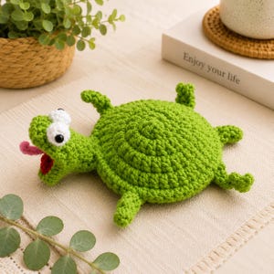 Op de afbeelding: Een felgroene gehaakte schildpad met een rond schild, vier kleine pootjes en een kop met grote witte ogen, zwarte pupillen en een open mond die een rode binnenkant en een uitstekende roze tong laat zien, gemaakt van textielgaren.