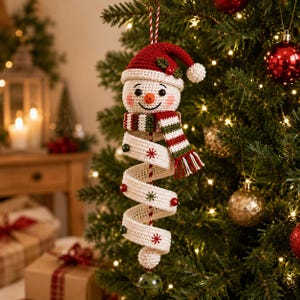 Könnte beinhalten: Ein gehäkelter Schneemann-Anhänger mit roter Mütze und Stechpalme, einem gestreiften Schal und einem langen, weißen spiralförmigen Körper, der mit kleinen roten und grünen gestickten Sternen und Perlen verziert ist und unten eine grüne Metallglocke trägt.