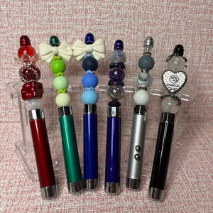 May include: Six pens with metallic barrels in red, green, blue, purple, silver, and black. Each pen features a decorative topper made of various beads, including silicone spheres, clear faceted crystals, white bows, and a white heart charm.
