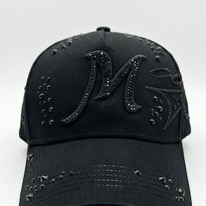 Puede incluir: Gorra de béisbol negra con visera curva. La gorra presenta una gran "M" adornada en la parte delantera, junto con tachuelas decorativas. La gorra está hecha de una tela texturizada y tiene un botón en la parte superior.