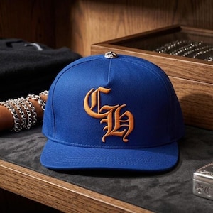 May include: A royal blue baseball cap with a flat brim. The cap features an orange embroidered logo on the front. A silver metal button is on top of the cap. The cap is displayed on a dark gray surface.
