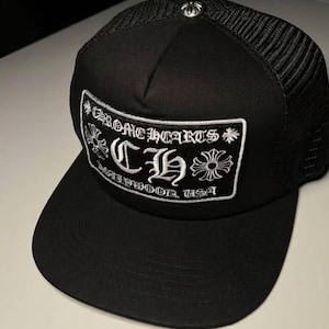 Puede incluir: Gorra de camionero negra con parte trasera de malla y visera curva. El panel frontal presenta un parche bordado blanco con el texto "CHROME HEARTS" y "MALIBU, USA". Un emblema plateado está en la parte superior.