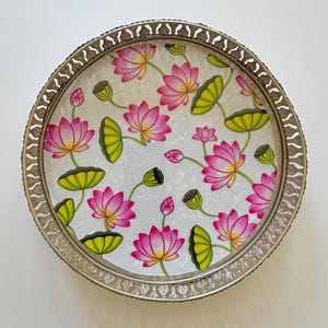 May include: A round serving tray with a metallic silver-toned rim featuring a decorative cut-out pattern. The interior surface displays a repeating pattern of pink lotus flowers, green lily pads, and closed flower buds on a white background.