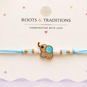 May include: A light blue string bracelet featuring a gold-toned elephant charm with a blue circular inlay and clear crystals. The bracelet is accented with small gold-toned beads, pale pink beads, and light blue beads on each side of the charm.