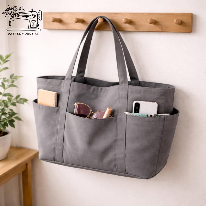May include: A gray tote bag with multiple exterior pockets, hanging on a wall. The pockets contain a notebook, sunglasses, lipstick, and a smartphone. The bag has two handles and the text "PATTERN MINT CO" with a sewing machine illustration.