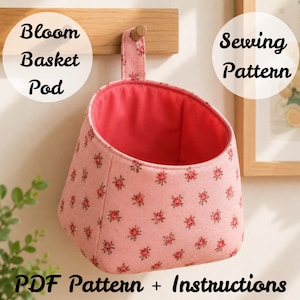 May include: A pink fabric Bloom Basket Pod with a floral pattern and a red interior. The pod is hanging from a wooden peg. The image also includes the text "Sewing Pattern" and "PDF Pattern + Instructions."