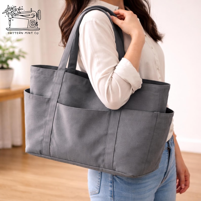 May include: A gray tote bag with multiple exterior pockets and sturdy handles. The bag is made of a durable fabric and features a rectangular shape. The bag is being held by a person wearing a white shirt and blue jeans. The bag has the text "PATTERN MINT CO" on it.