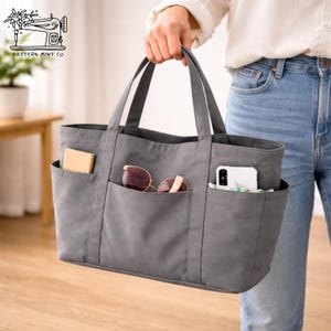 May include: A gray tote bag with multiple exterior pockets, held by a person. The pockets contain a small box, sunglasses, and a smartphone. The bag has two handles and is made of a sturdy fabric.