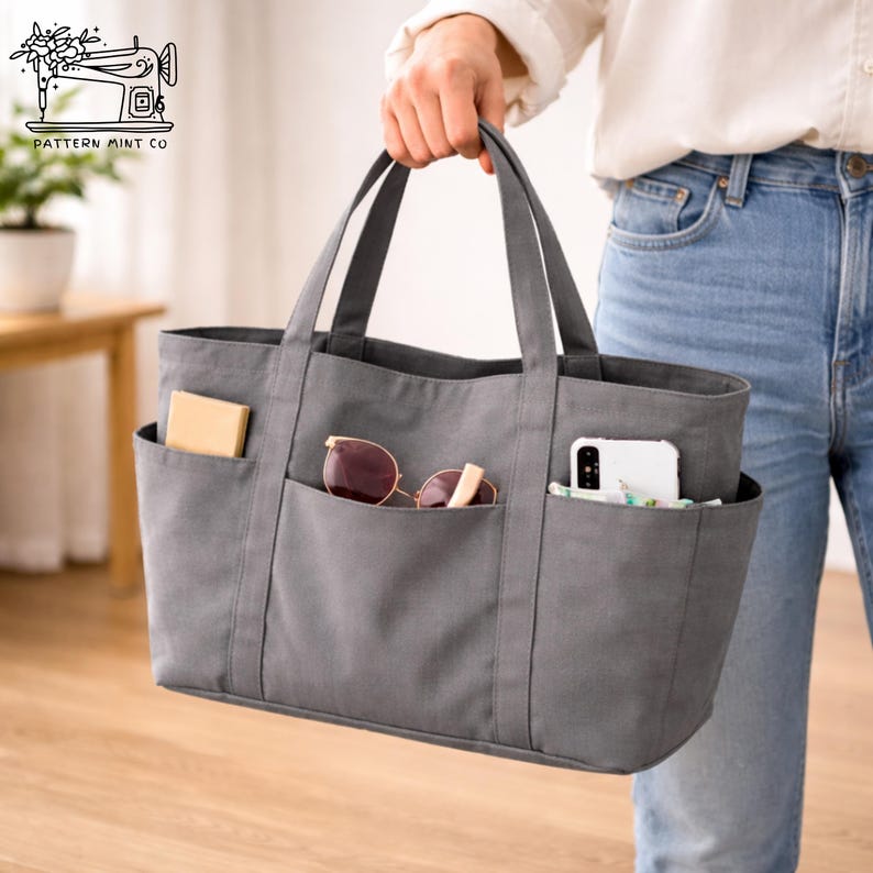 May include: A gray tote bag with multiple exterior pockets, held by a person. The pockets contain a small box, sunglasses, and a smartphone. The bag has two handles and is made of a sturdy fabric.