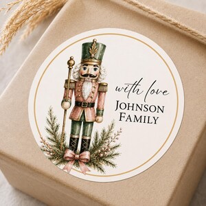 May include: A round sticker featuring a watercolour illustration of a nutcracker in a green and pink uniform, standing amidst pine branches and a pink bow. The text reads with love JOHNSON FAMILY in black script and serif fonts on a cream background.