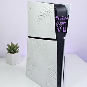 Pode incluir: Um conjunto de placas frontais brancas personalizadas para PS5 com padrões de folhas de palmeira em relevo e um algarismo romano VI grande. O centro preto da consola exibe um logótipo roxo e preto State of Leonida VI. Texto: CUSTOM LEONIDA PS5.