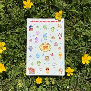 May include: A rectangular sticker sheet titled ANIMAL CROSSING EDITION featuring various colorful cartoon characters, fruits, and icons. In the center, a logo reads TINY TOMATO CO. with an orange cat illustration. The sheet has a white background.