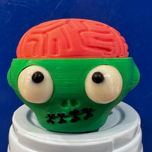 Peut inclure: Une tête de zombie imprimée en 3D avec un cerveau rouge et un visage vert. Le zombie a de grands yeux blancs avec des pupilles noires et une bouche cousue. La tête est posée sur une surface blanche sur fond bleu.