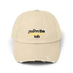 May include: A distressed beige baseball cap with the text "pull on the yellow tab" in black and yellow. The cap has a curved brim and a worn, vintage look, with distressed edges.