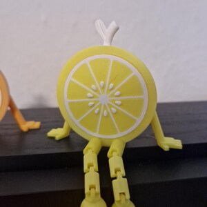 May include: A whimsical, yellow lemon slice character with articulated legs and arms, and a white rind detail. The lemon has a white stem and is sitting on a dark surface. The design is playful and cartoonish.