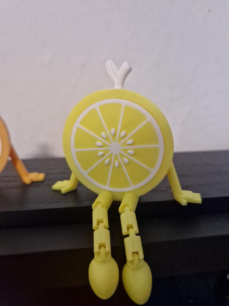 May include: A whimsical, yellow lemon slice character with articulated legs and arms, and a white rind detail. The lemon has a white stem and is sitting on a dark surface. The design is playful and cartoonish.