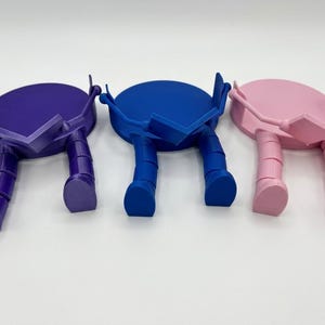 May include: Three colourful phone stands in purple, blue, and pink. Each stand has a circular top with a rectangular support and two legs, giving them a playful, robot-like appearance. The stands are set against a white background.