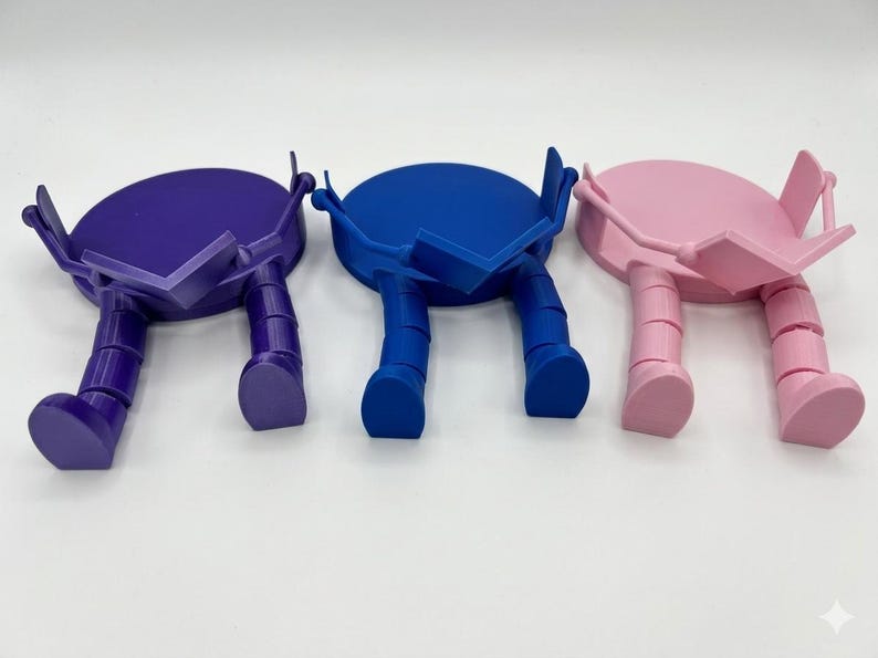 May include: Three colourful phone stands in purple, blue, and pink. Each stand has a circular top with a rectangular support and two legs, giving them a playful, robot-like appearance. The stands are set against a white background.