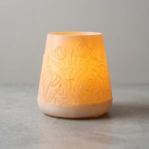 May include: A glowing, off-white candle holder with a floral tulip design. The holder has a wide base and a slightly tapered shape, with the tulip design carved into the surface. The light inside casts a warm glow, highlighting the intricate details of the flowers.