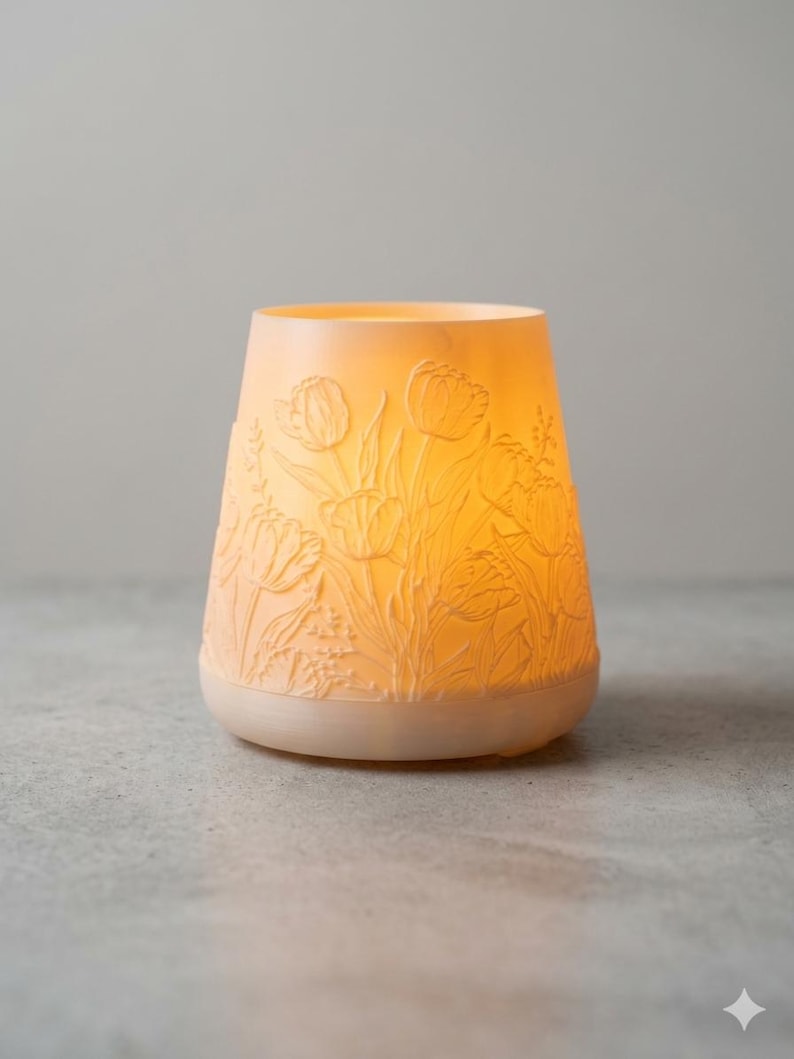 May include: A glowing, off-white candle holder with a floral tulip design. The holder has a wide base and a slightly tapered shape, with the tulip design carved into the surface. The light inside casts a warm glow, highlighting the intricate details of the flowers.