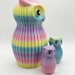 Puede incluir: Colecci&oacute;n de tres figuras de b&uacute;hos de diferentes tama&ntilde;os. El b&uacute;ho m&aacute;s grande es un arco&iacute;ris de colores, mientras que los m&aacute;s peque&ntilde;os son tonos pastel de morado y azul. Todos tienen una textura acanalada y ojos grandes y redondos.
