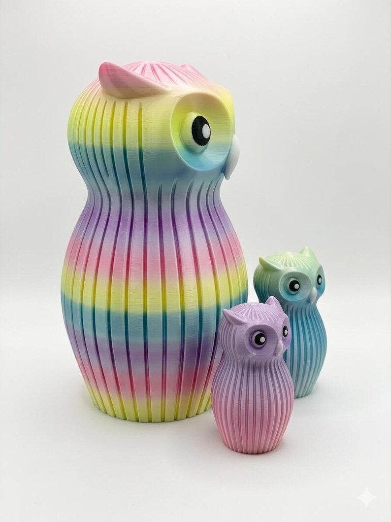Puede incluir: Colecci&oacute;n de tres figuras de b&uacute;hos de diferentes tama&ntilde;os. El b&uacute;ho m&aacute;s grande es un arco&iacute;ris de colores, mientras que los m&aacute;s peque&ntilde;os son tonos pastel de morado y azul. Todos tienen una textura acanalada y ojos grandes y redondos.