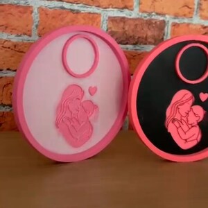 Op de afbeelding: Twee ronde onderzetters met roze en zwarte randen. Elke onderzetter heeft een roze silhouet van een moeder en kind, met een hartdetail. De achtergrond is een bakstenen muur.