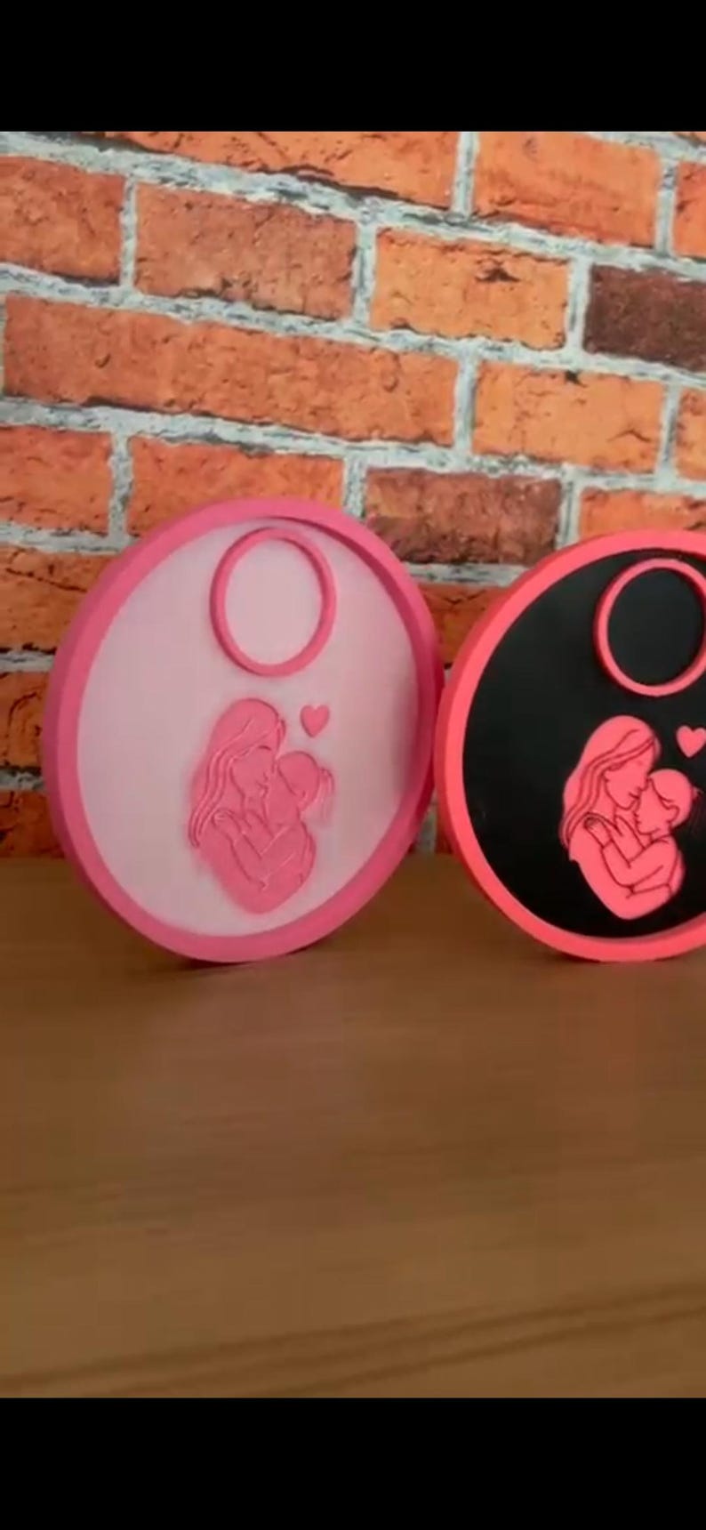 Op de afbeelding: Twee ronde onderzetters met roze en zwarte randen. Elke onderzetter heeft een roze silhouet van een moeder en kind, met een hartdetail. De achtergrond is een bakstenen muur.